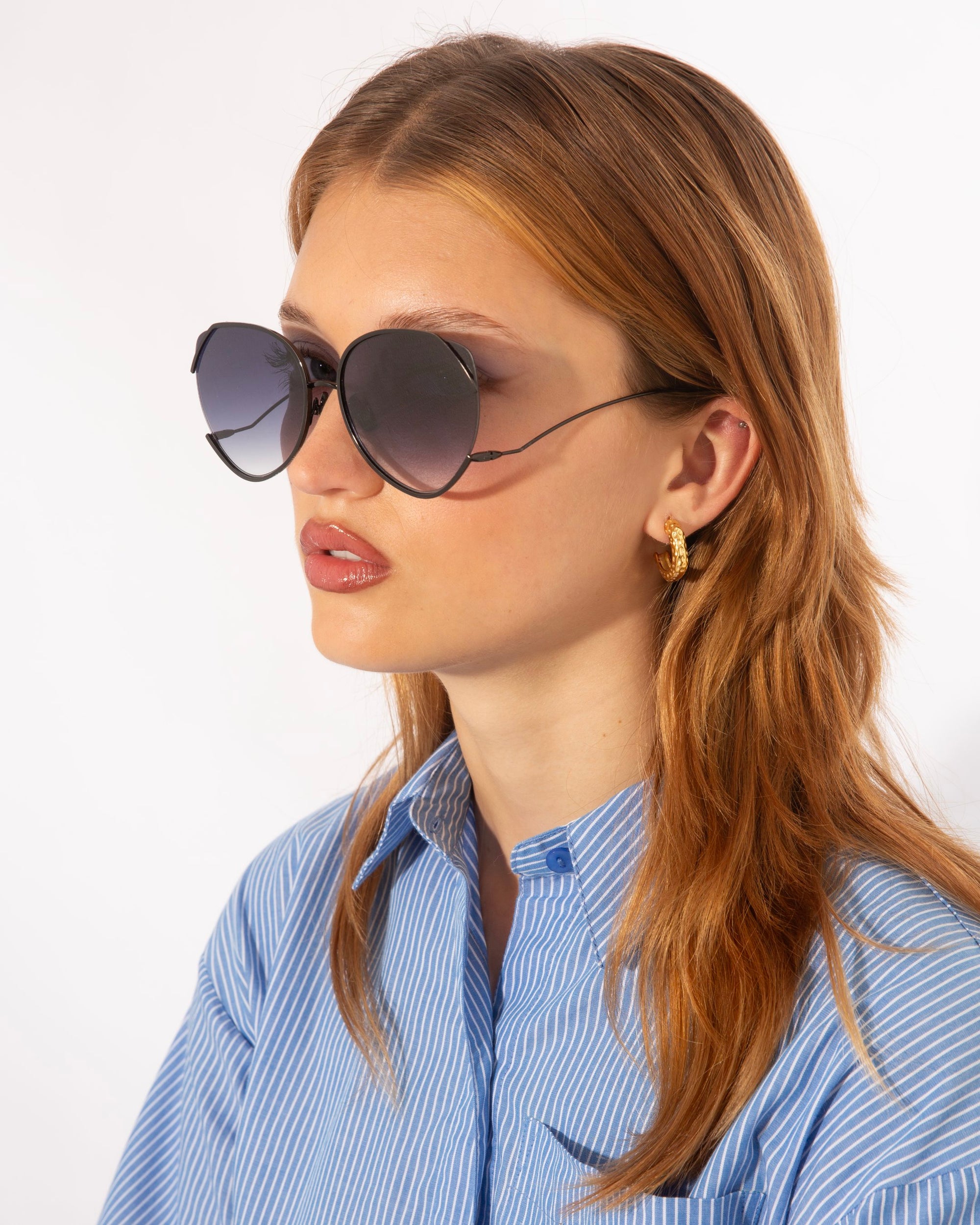 A person with long, straight, reddish hair is wearing oversized For Art's Sake® Wonderland sunglasses with 100% UV protection and a blue and white striped shirt. They also have gold hoop earrings and a serious expression, standing against a plain, white background.