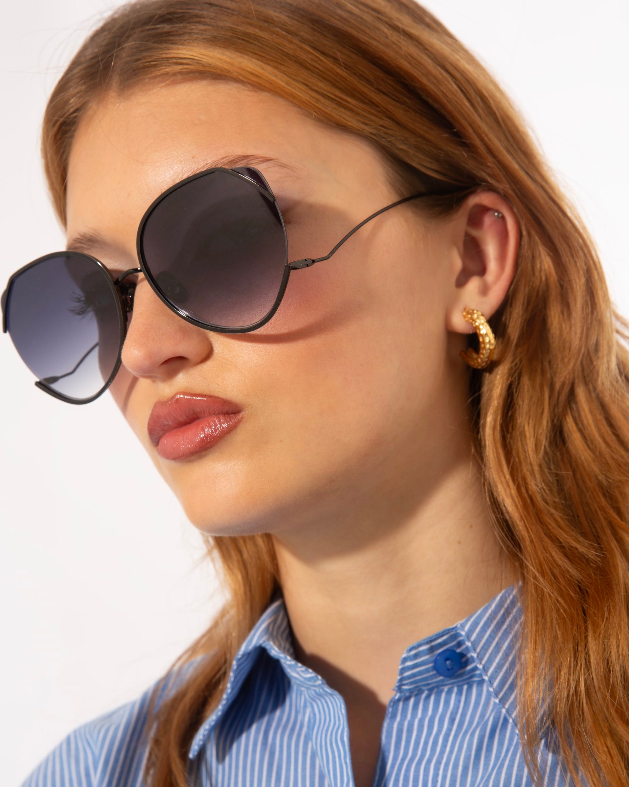 A person with long, light brown hair wears large, round For Art's Sake® Wonderland sunglasses with jadestone nosepads and UV protection. They sport a blue striped shirt with the top button undone and gold hoop earrings, looking to their left against a plain background.