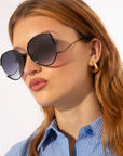 A person with long, light brown hair wears large, round For Art's Sake® Wonderland sunglasses with jadestone nosepads and UV protection. They sport a blue striped shirt with the top button undone and gold hoop earrings, looking to their left against a plain background.