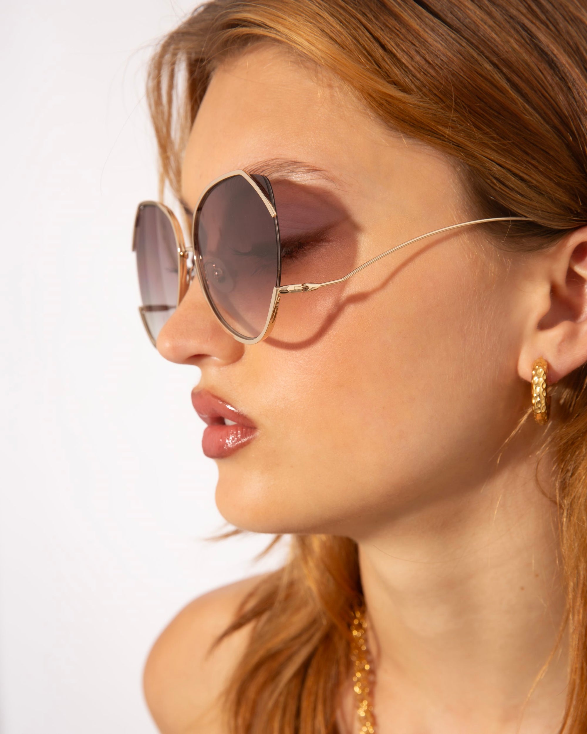 A young woman with light brown hair wearing large, round For Art&#39;s Sake® Wonderland sunglasses featuring jadestone nose pads and gold frames. She sports small gold hoop earrings. The lighting highlights her clear skin and neutral makeup, and she appears against a plain white background.