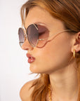 A young woman with light brown hair wearing large, round For Art's Sake® Wonderland sunglasses featuring jadestone nose pads and gold frames. She sports small gold hoop earrings. The lighting highlights her clear skin and neutral makeup, and she appears against a plain white background.