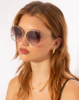 A woman with shoulder-length light brown hair is wearing large, round For Art's Sake® Wonderland sunglasses with thin gold frames and jadestone nosepads. She has gold hoop earrings and a gold chain necklace adorned with pearl accents. She is dressed in a black top, posed against a plain, light background.