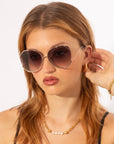 A young woman with long auburn hair is gazing forward while wearing large, round For Art's Sake® Wonderland sunglasses with ultralightweight lenses. She holds one hand up to her ear, displaying her gold earrings. She is also wearing a gold necklace with pearl accents and a gold bracelet. She has a neutral expression.