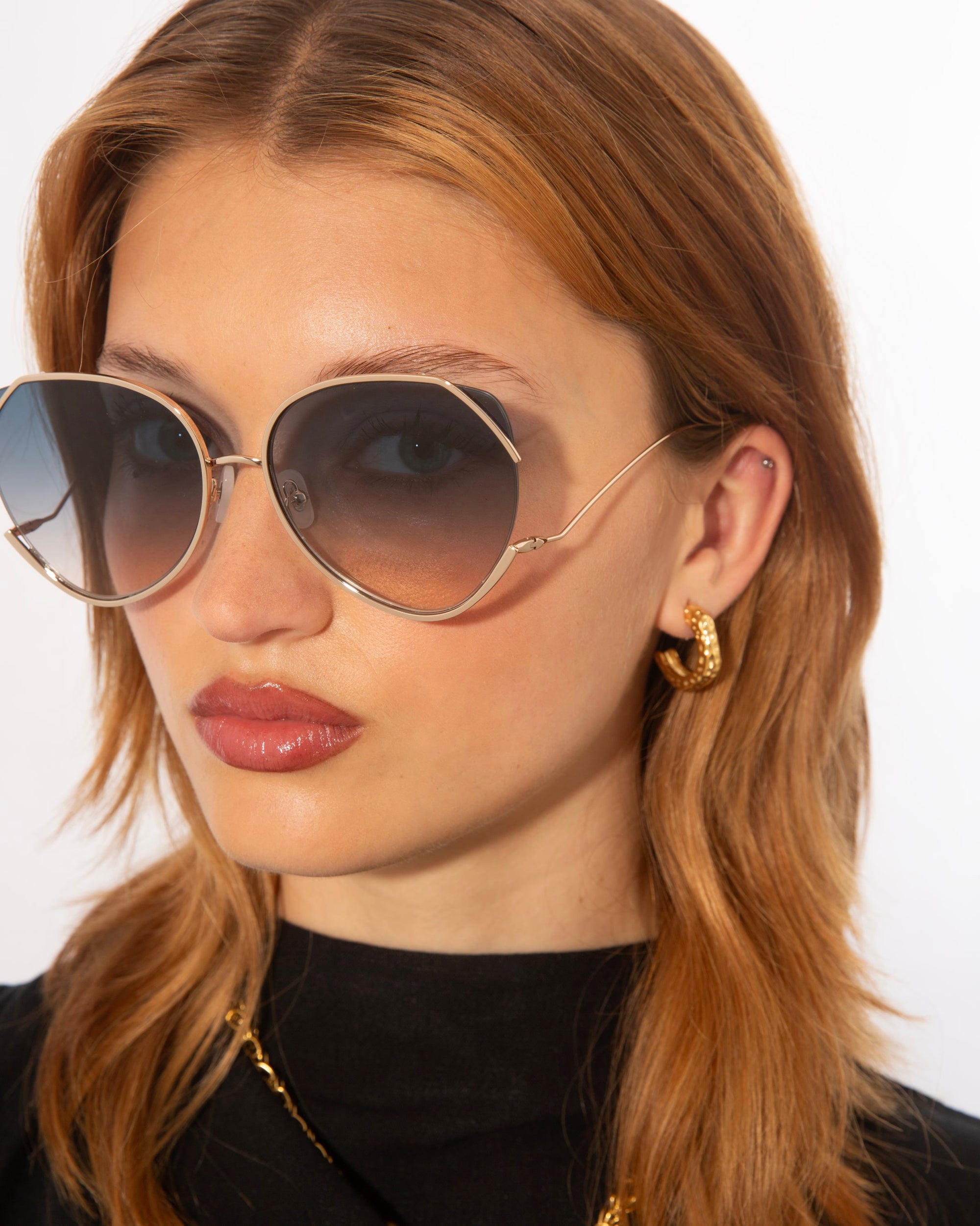 A person with long, light brown hair is wearing large, round For Art&#39;s Sake® Wonderland sunglasses featuring ultralightweight shatter-resistant lenses and gold trim. They have small gold hoop earrings and are dressed in a black top. The background is plain white.