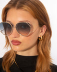 A person with long, light brown hair is wearing large, round For Art's Sake® Wonderland sunglasses featuring ultralightweight shatter-resistant lenses and gold trim. They have small gold hoop earrings and are dressed in a black top. The background is plain white.