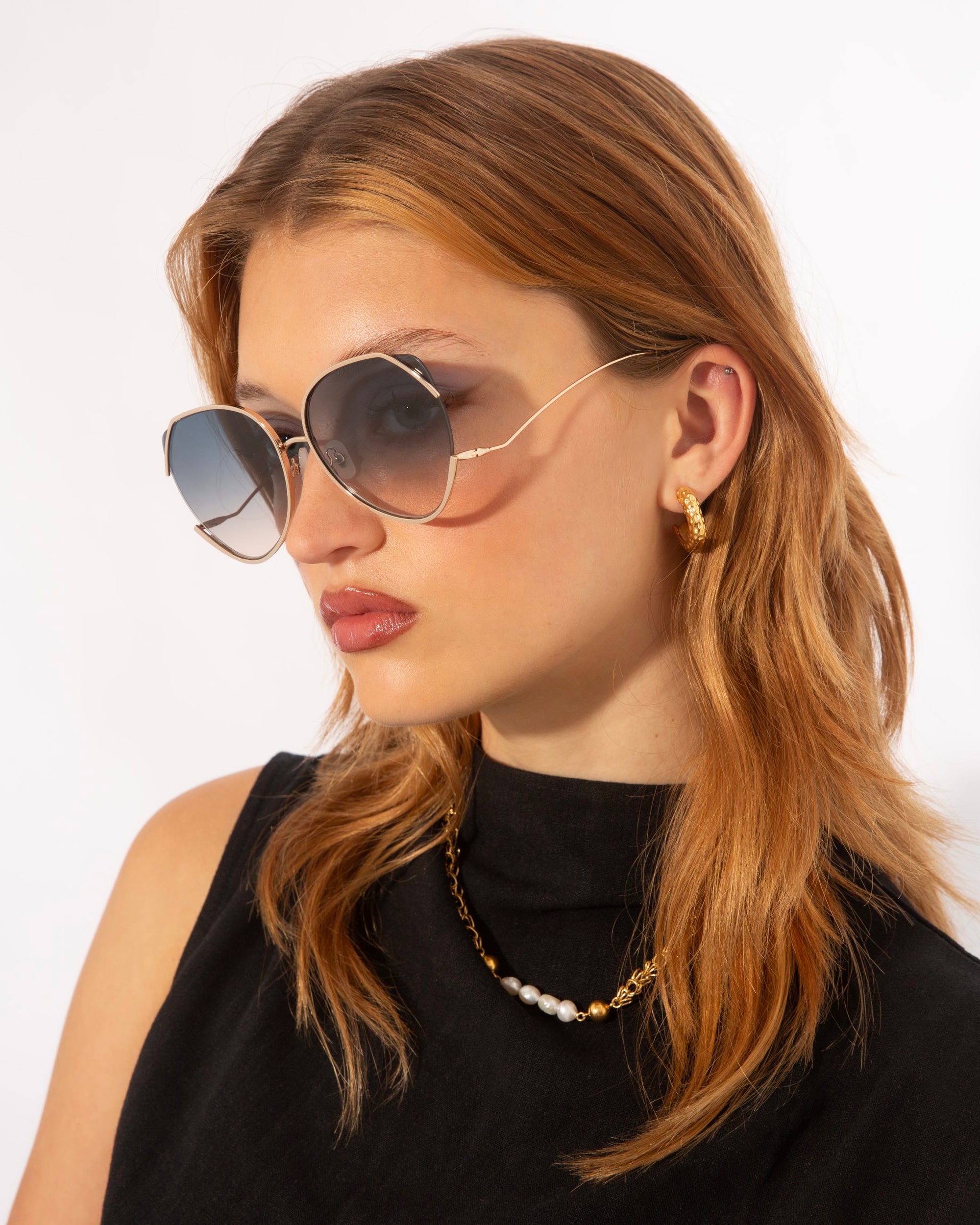 A woman with medium-length, light brown hair is wearing large, round For Art&#39;s Sake® Wonderland sunglasses with adjustable nosepads and 100% UV protection. She has a serious expression and is accessorized with gold hoop earrings and a gold necklace with small white beads. Her sleeveless black top stands out against the plain white background.