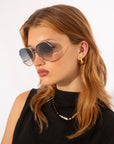 A woman with medium-length, light brown hair is wearing large, round For Art's Sake® Wonderland sunglasses with adjustable nosepads and 100% UV protection. She has a serious expression and is accessorized with gold hoop earrings and a gold necklace with small white beads. Her sleeveless black top stands out against the plain white background.