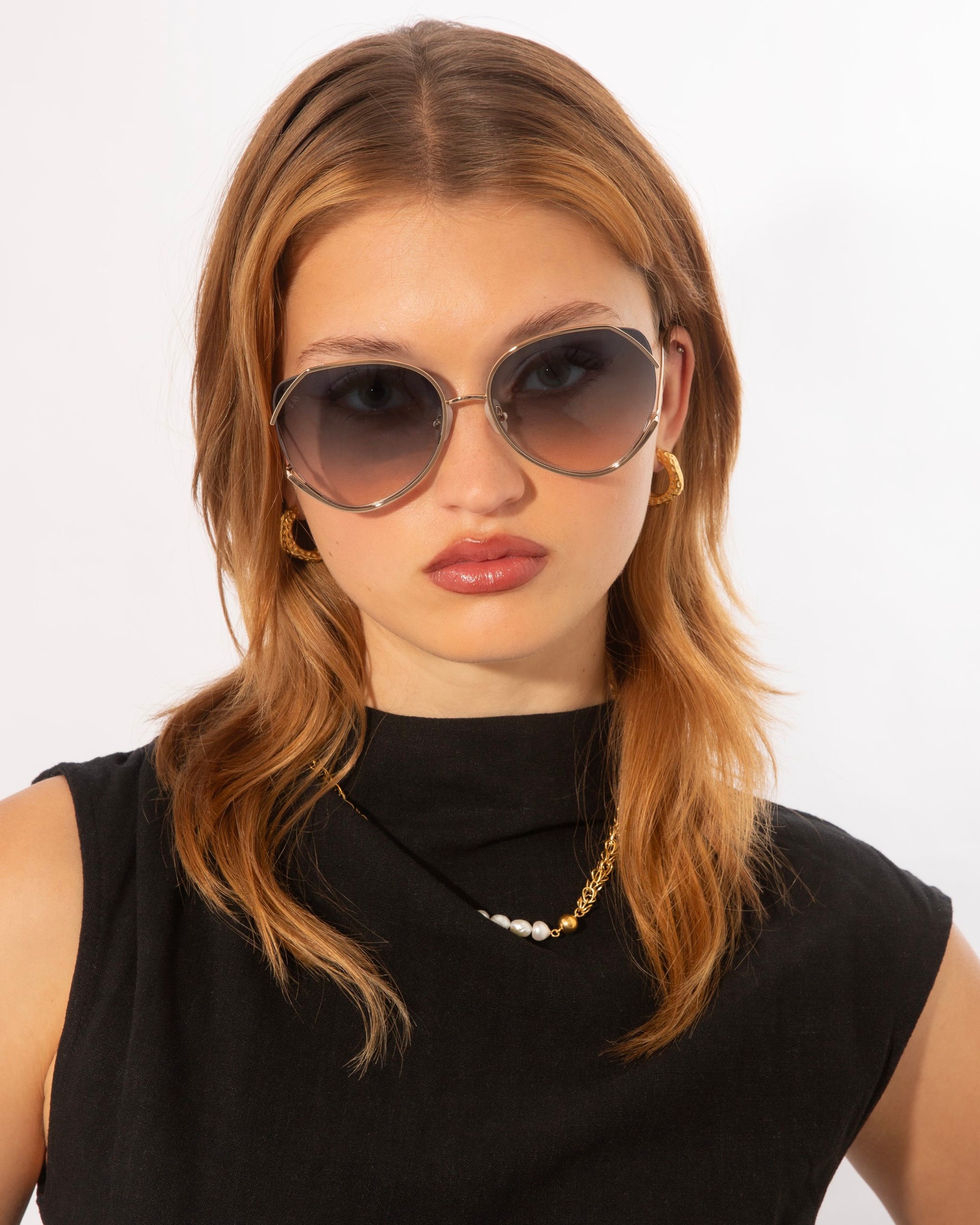 A woman with shoulder-length light brown hair is wearing large, round, dark sunglasses that offer 100% UV protection. She has a neutral expression and is dressed in a sleeveless black top with a gold necklace featuring a single white pearl. Her golden hoop earrings complement the ultralightweight design of her Wonderland by For Art&#39;s Sake® eyewear. The background is white.
