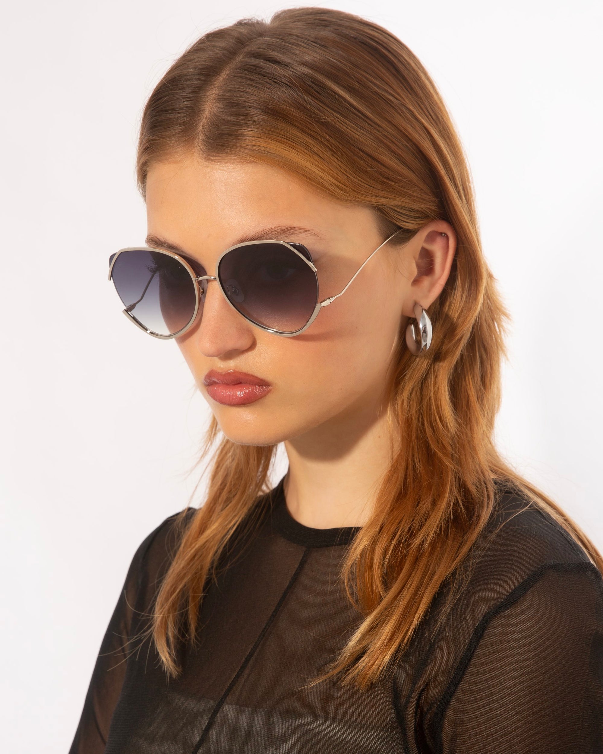 A woman with reddish-brown hair is wearing large, hexagonal For Art&#39;s Sake® Wonderland sunglasses featuring jadestone nose pads and offering 100% UV protection. She has hoop earrings and is dressed in a sheer black top with a solid black panel covering her torso. The background is plain white.