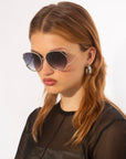 A woman with reddish-brown hair is wearing large, hexagonal For Art's Sake® Wonderland sunglasses featuring jadestone nose pads and offering 100% UV protection. She has hoop earrings and is dressed in a sheer black top with a solid black panel covering her torso. The background is plain white.