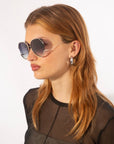 A woman with long, light red hair wears large, round For Art's Sake® Wonderland sunglasses with 100% UV protection and jadestone nose pads. She is dressed in a sheer black top and is gazing to the side against a plain white background.