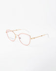 A pair of eyeglasses with gold-colored, thin, metal frames and pink accents around the lenses. The temples feature a decorative pattern and clear, adjustable nose pads. These 18-karat gold-plated glasses are displayed against a plain white background. They are the Julie by For Art's Sake®.