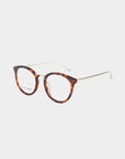 The Jackie by For Art's Sake® features a pair of round, tortoiseshell eyeglasses with thin, gold-colored metal temples and clear lenses. The structure combines a classic brown and amber pattern on the frame with sleek metal arms, creating a sophisticated and stylish appearance. Available with blue light filter for enhanced protection.