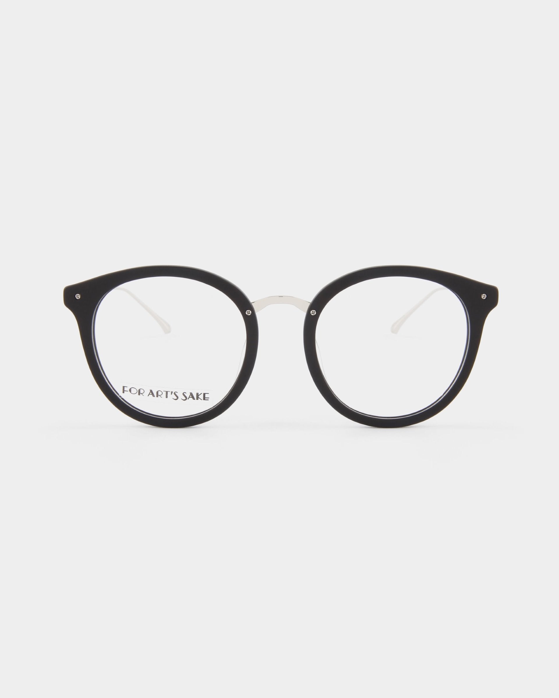 A pair of round, black-rimmed eyeglasses with clear lenses, displayed against a white background. The inside of the left temple arm is inscribed with &quot;FOR ARTS SAKE.&quot; The glasses feature UV protection lenses and have a classic and minimalistic design. These are the Jackie by For Art&#39;s Sake®.