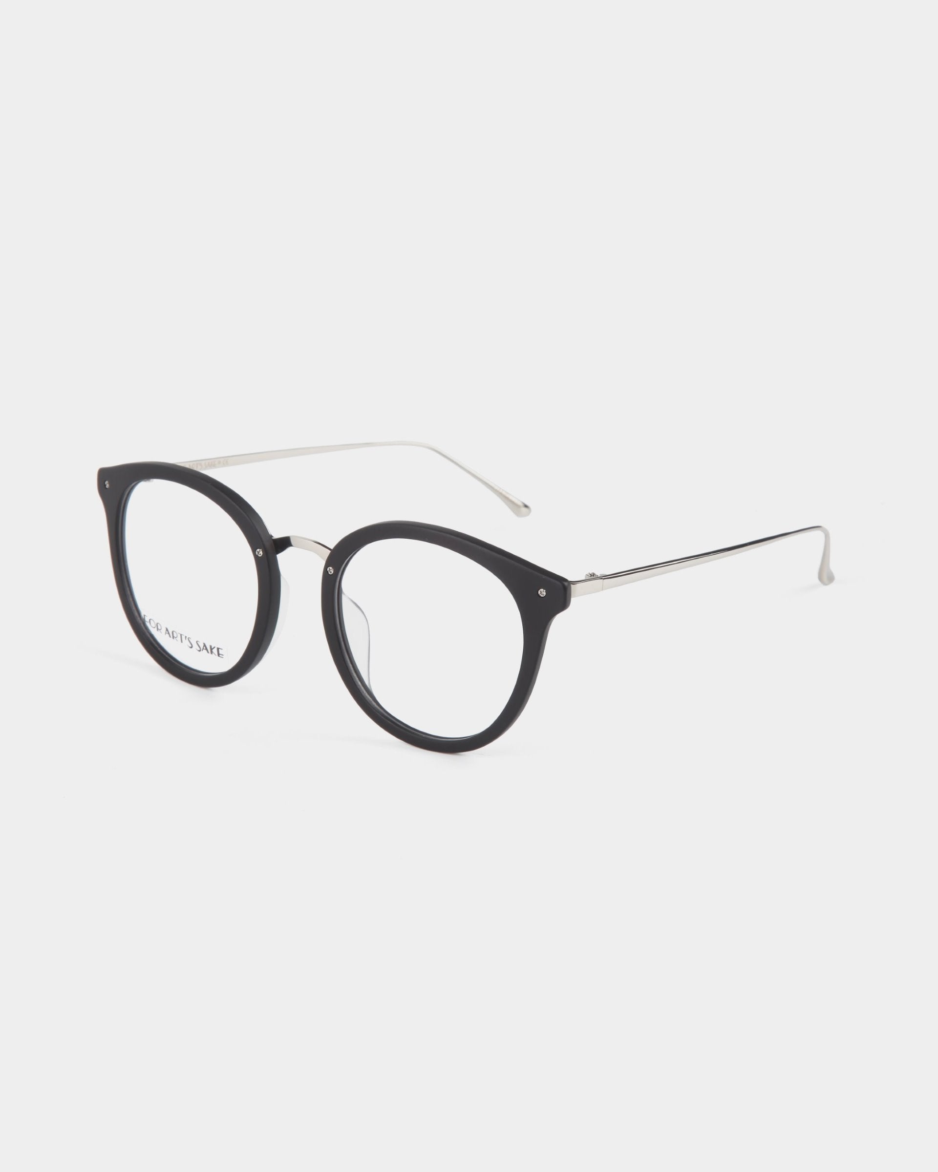 A pair of Jackie eyeglasses by For Art&#39;s Sake® with round black frames and thin metal temples. The lenses are clear and feature blue light filters, making the design modern and minimalist. The eyeglasses are displayed on a white background.