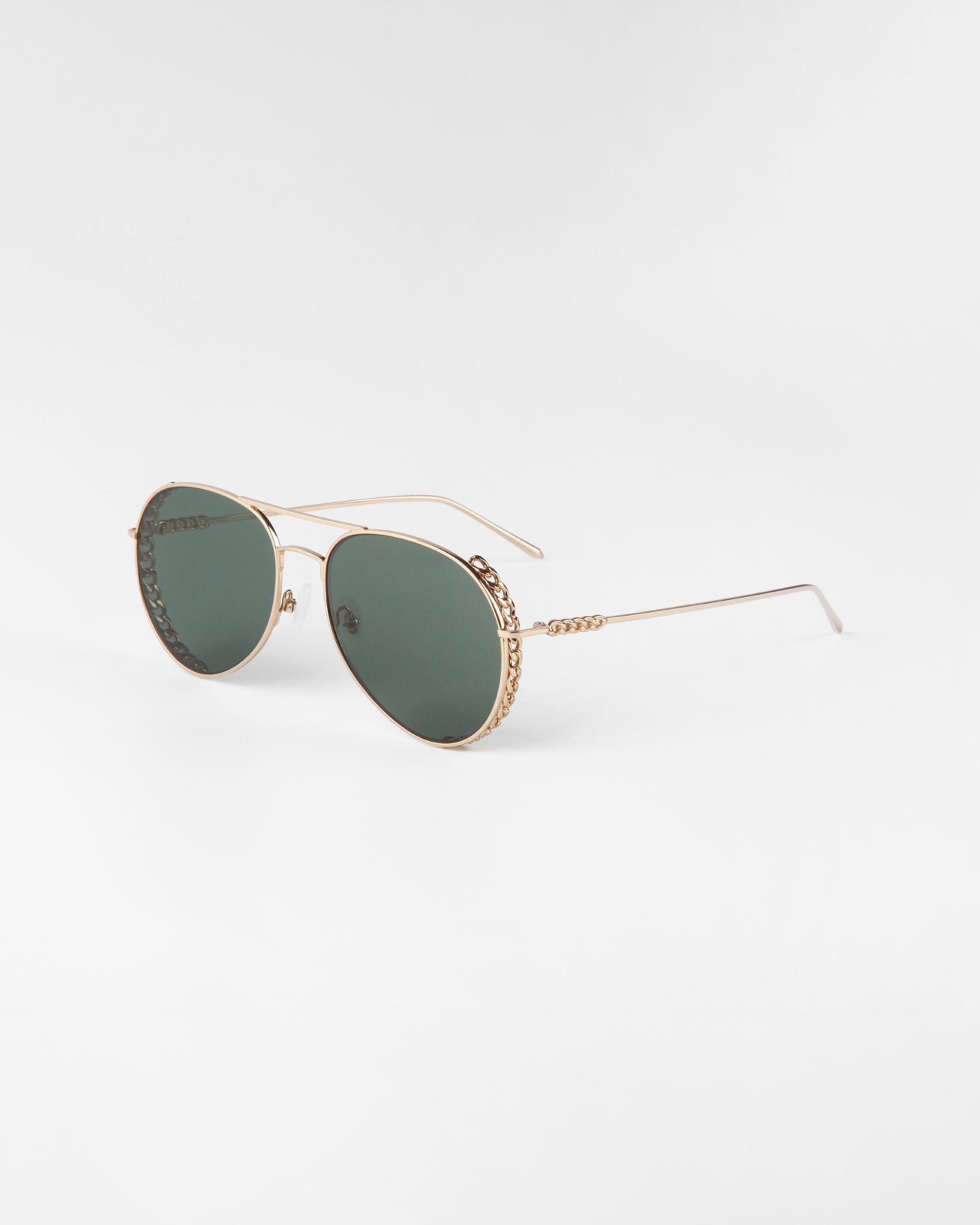 A pair of stylish Links aviator sunglasses by For Art&#39;s Sake® with thin gold-plated stainless steel frames and green nylon lenses. The gold temples feature a subtle chain detail near the hinges, adding a touch of elegance to the classic design. The Links sunglasses are set against a plain, light background.