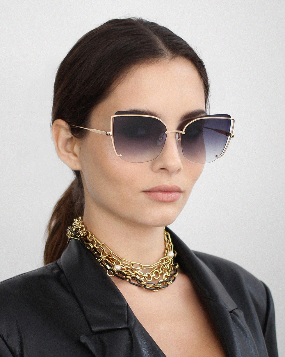 A woman wearing large, square, gradient Meow sunglasses by For Art&#39;s Sake® with adjustable nosepads, a chunky 18-karat gold plated chain necklace, and a black faux leather blazer with her hair tied back in a ponytail. She is facing forward with a neutral expression against a plain, light background.