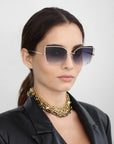 A woman wearing large, square, gradient Meow sunglasses by For Art's Sake® with adjustable nosepads, a chunky 18-karat gold plated chain necklace, and a black faux leather blazer with her hair tied back in a ponytail. She is facing forward with a neutral expression against a plain, light background.