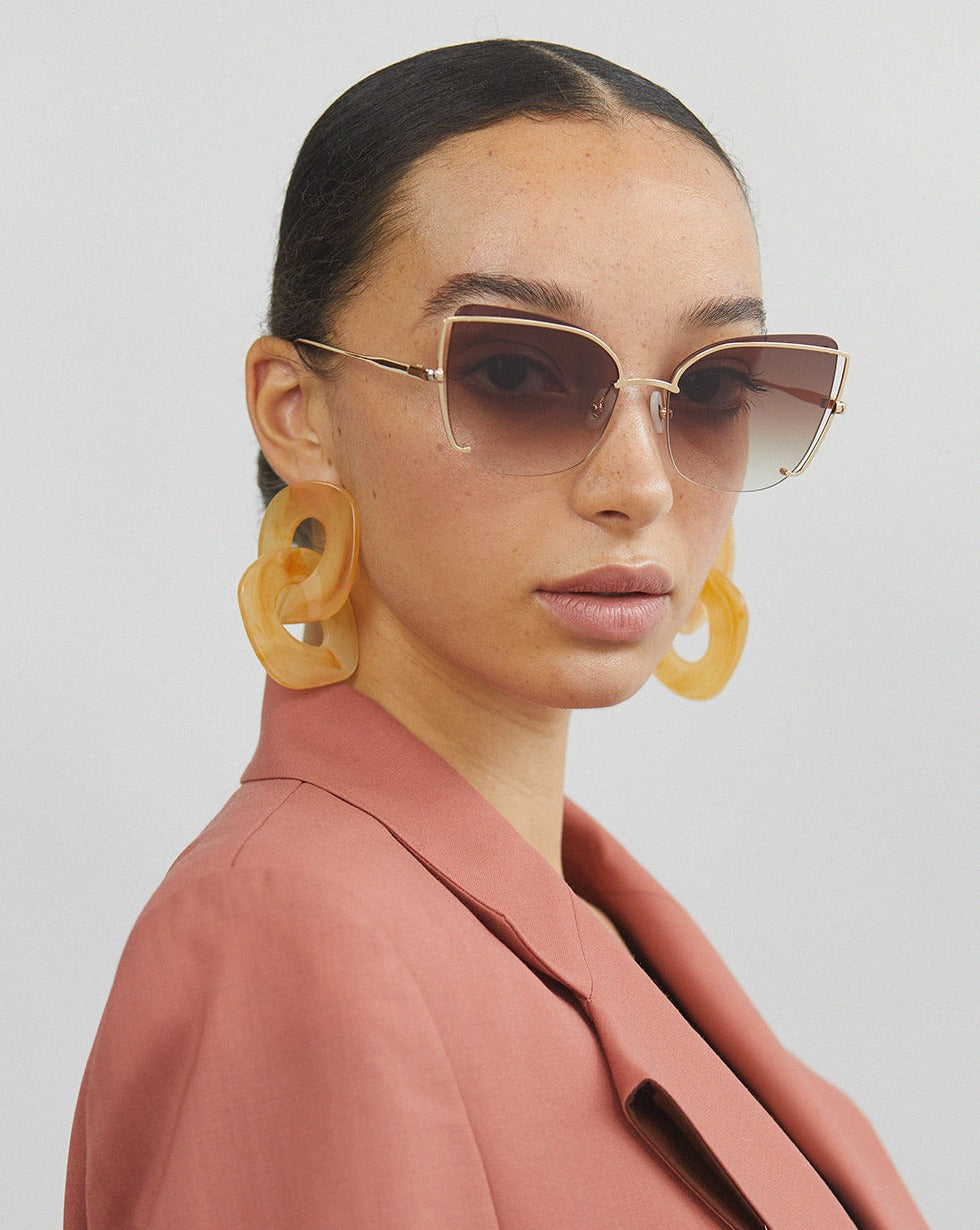 A person with neatly pulled-back hair wears large, angular Meow sunglasses by For Art's Sake® featuring adjustable nosepads, oversized yellow earrings, and a peach-colored blazer. The background is plain, and the person's expression is neutral.