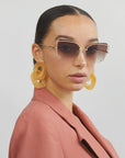 A person with neatly pulled-back hair wears large, angular Meow sunglasses by For Art's Sake® featuring adjustable nosepads, oversized yellow earrings, and a peach-colored blazer. The background is plain, and the person's expression is neutral.