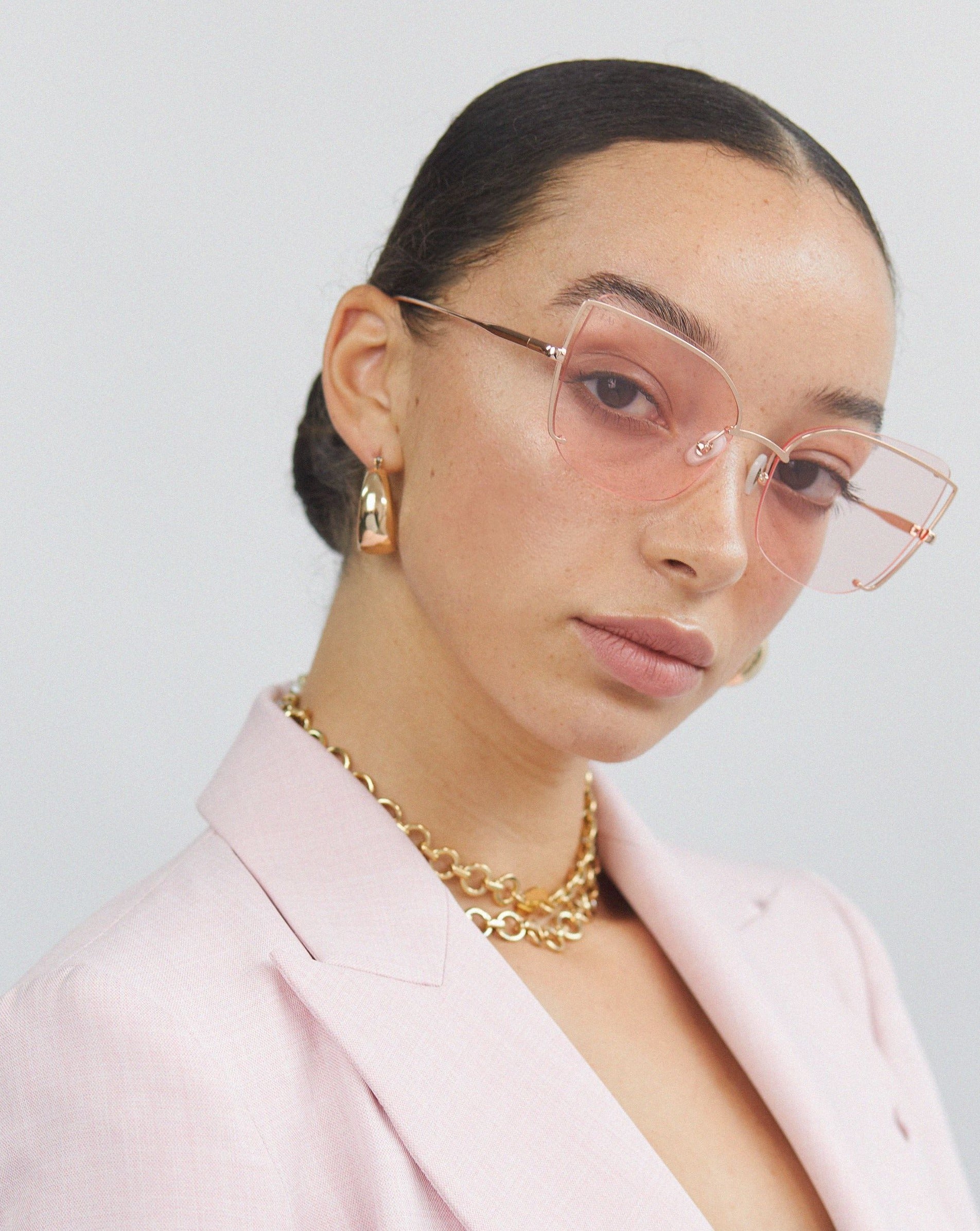 A person with slicked-back hair is wearing For Art&#39;s Sake® Meow - Champagne sunglasses, which feature a large cat-eye silhouette with pink-tinted nylon lenses and gold plated stainless steel frames. Gold earrings and a chunky gold chain necklace complete the look. They are dressed in a light pink blazer over a black top, posing against a plain, light background.