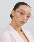 A person with slicked-back hair is wearing For Art's Sake® Meow - Champagne sunglasses, which feature a large cat-eye silhouette with pink-tinted nylon lenses and gold plated stainless steel frames. Gold earrings and a chunky gold chain necklace complete the look. They are dressed in a light pink blazer over a black top, posing against a plain, light background.