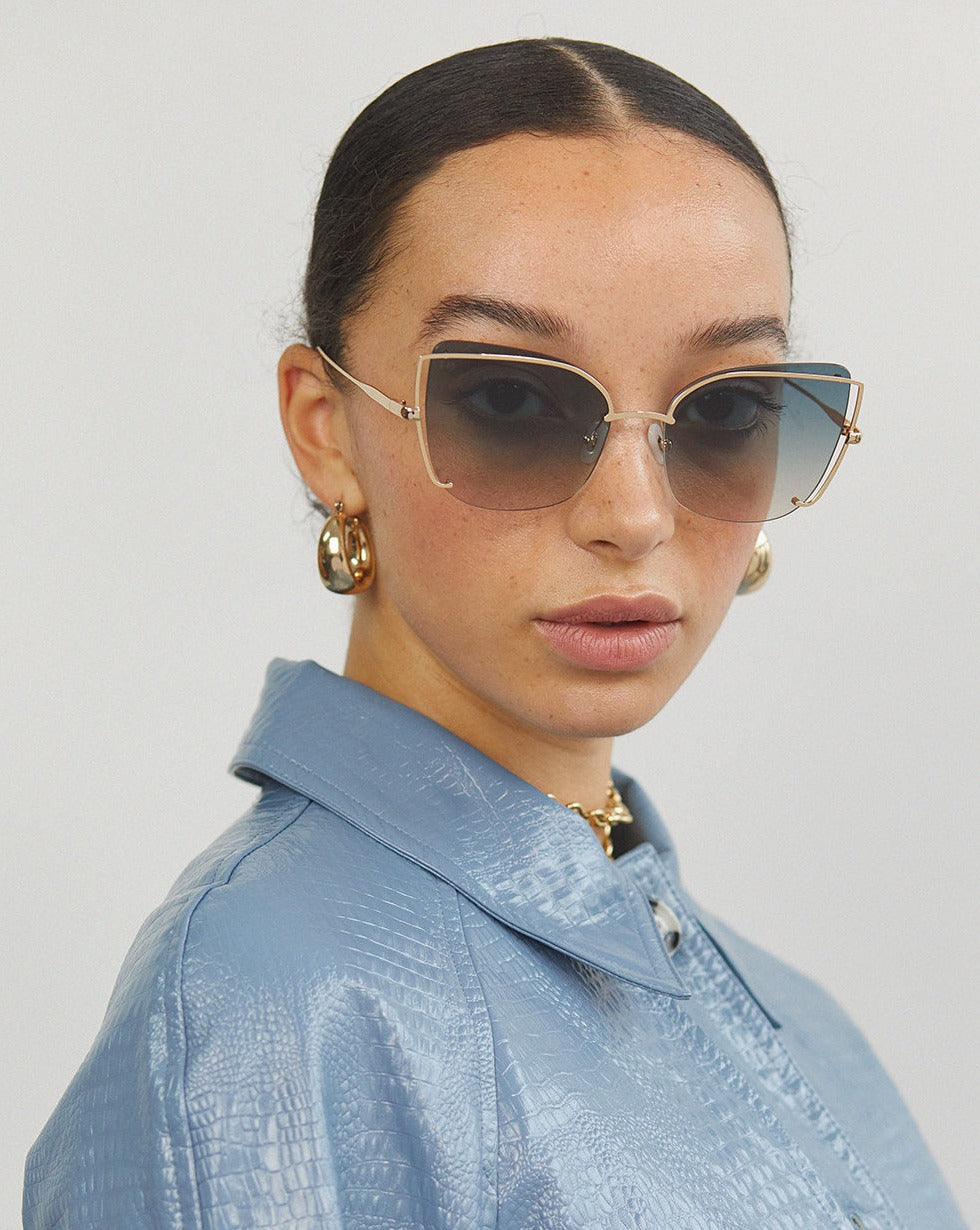 Wearing For Art&#39;s Sake® Meow shades and gold hoop earrings, the individual sports a light blue textured jacket. Their dark hair is slicked back into a low bun, and they gaze directly at the camera against a plain, light-colored background.