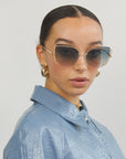 Wearing For Art's Sake® Meow shades and gold hoop earrings, the individual sports a light blue textured jacket. Their dark hair is slicked back into a low bun, and they gaze directly at the camera against a plain, light-colored background.