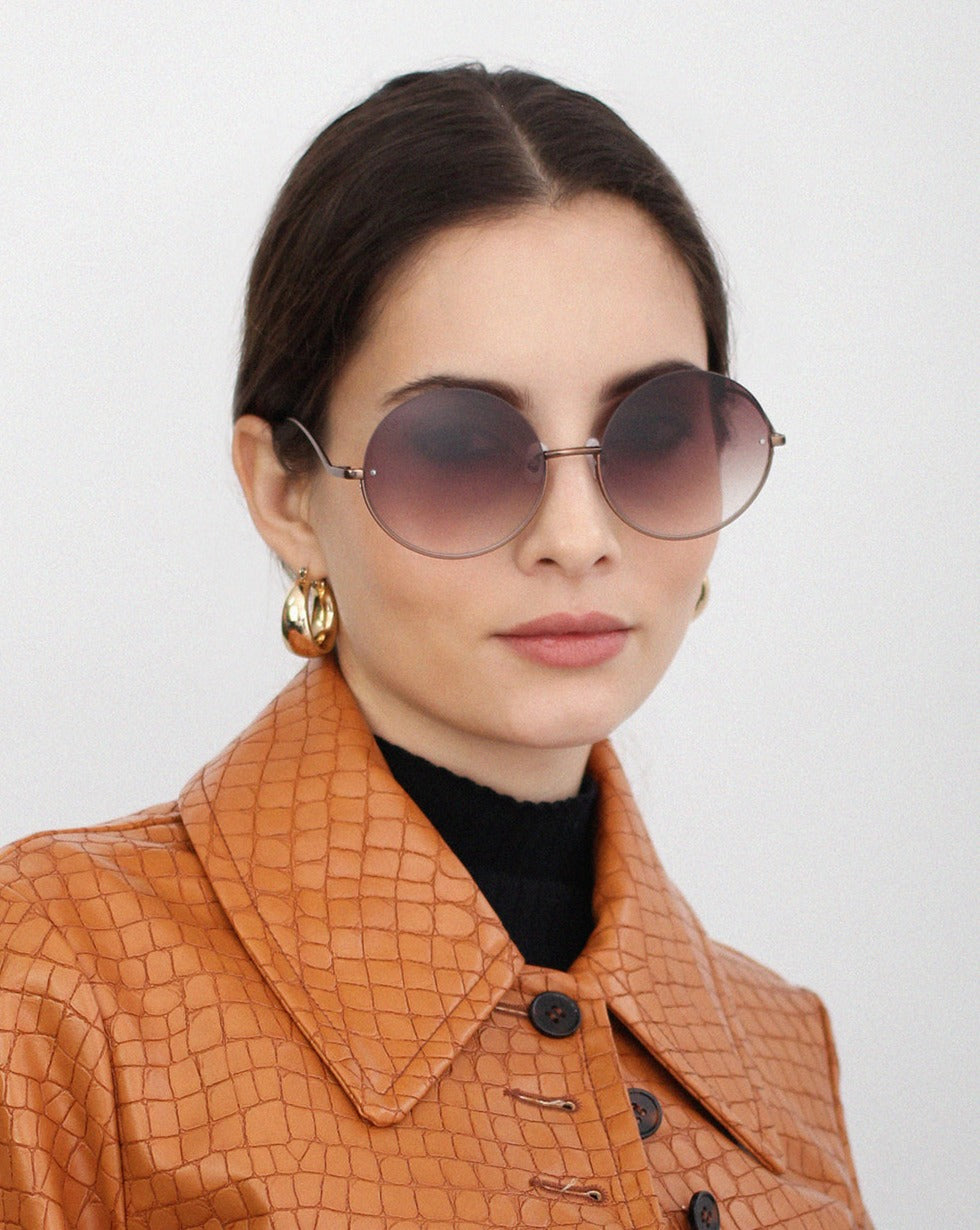 A person with dark hair is wearing round, gradient sunglasses called Oceana by For Art's Sake® with stainless steel frames and UVA & UVB-protected nylon lenses. They have a neutral expression and their hair is pulled back, while sporting a light brown, textured coat. The background is plain and light-colored.
