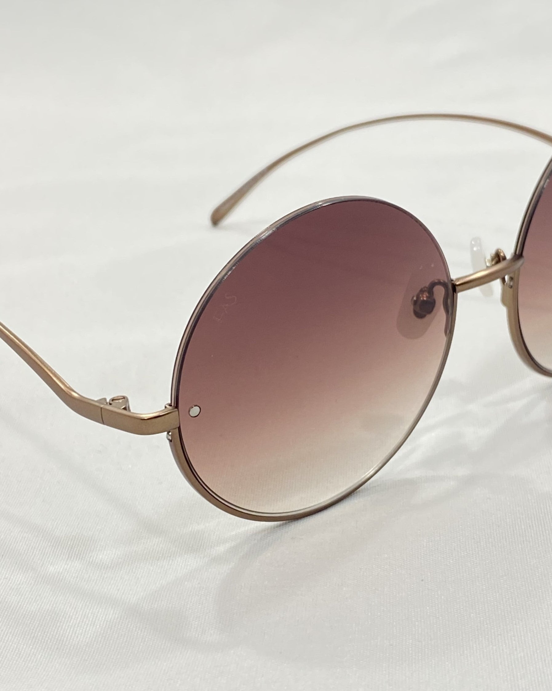 A pair of Oceana frames by For Art&#39;s Sake®, featuring a rounded design with thin gold accents and gradient lenses that transition from pink at the top to clear at the bottom. Perfect for a throw-on-and-go look, these sunglasses rest on a white surface, showcasing a small visible engraving near the right edge.