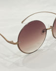 A pair of Oceana frames by For Art's Sake®, featuring a rounded design with thin gold accents and gradient lenses that transition from pink at the top to clear at the bottom. Perfect for a throw-on-and-go look, these sunglasses rest on a white surface, showcasing a small visible engraving near the right edge.