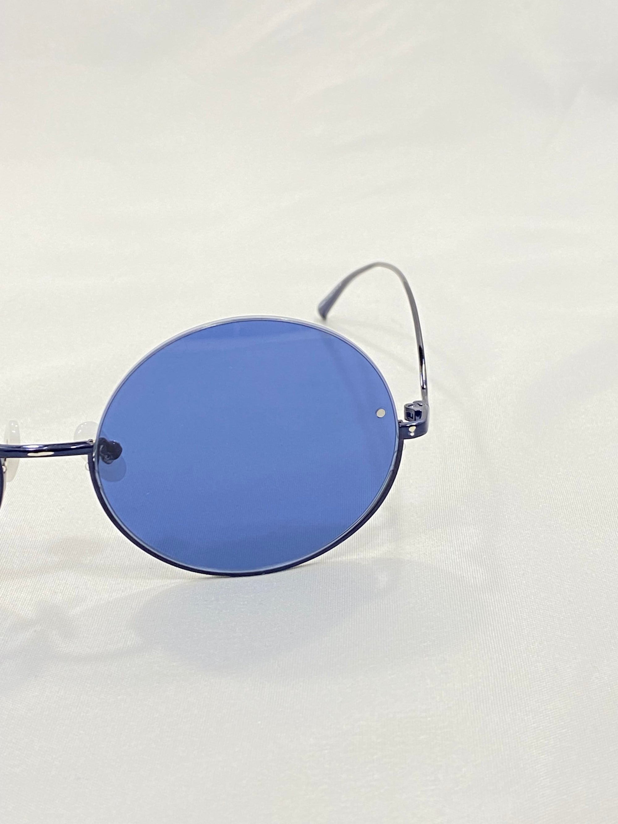 A close-up image of a pair of Oceana sunglasses by For Art&#39;s Sake® with blue-tinted lenses. The sunglasses feature thin metal frames and sport a pared-back shape, set against a plain, light-colored background. The left lens and side arm are prominently visible. Enjoy free shipping on your purchase!