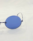 A close-up image of a pair of Oceana sunglasses by For Art's Sake® with blue-tinted lenses. The sunglasses feature thin metal frames and sport a pared-back shape, set against a plain, light-colored background. The left lens and side arm are prominently visible. Enjoy free shipping on your purchase!