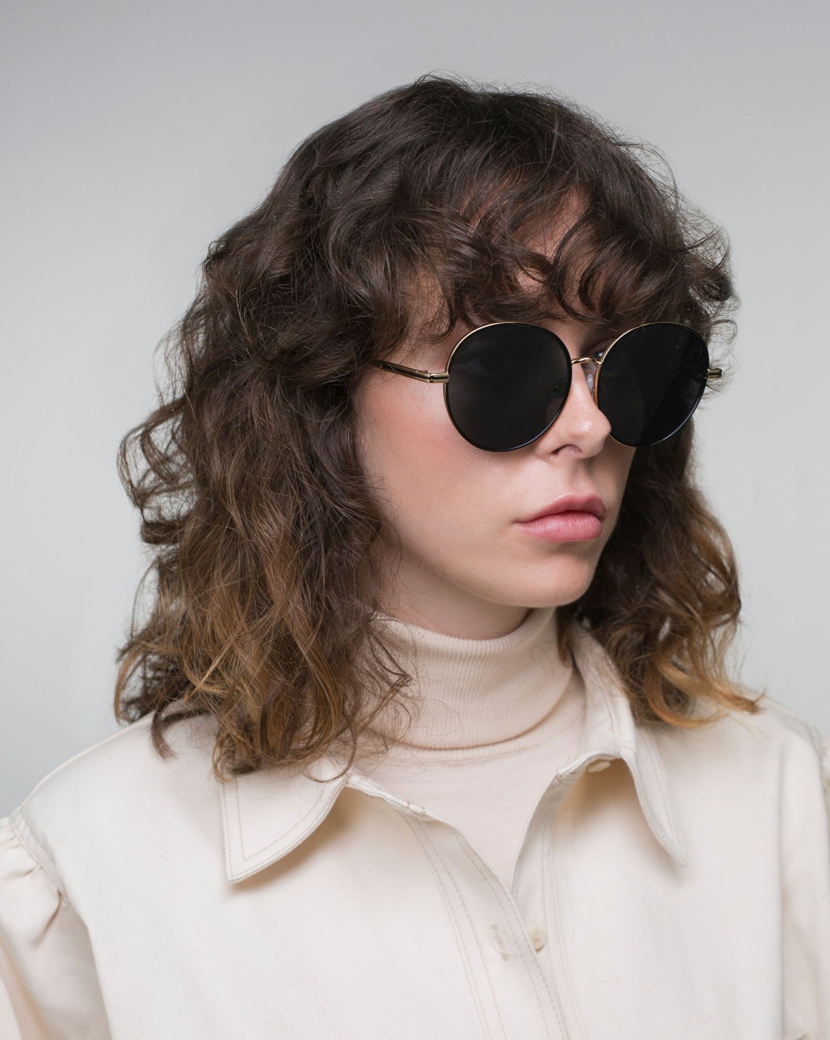 A person with shoulder-length curly hair and bangs is wearing the &quot;Lemon&quot; round dark sunglasses from For Art&#39;s Sake® with lightweight nylon lenses, paired with a light-colored turtleneck sweater, posing against a plain, light gray background.