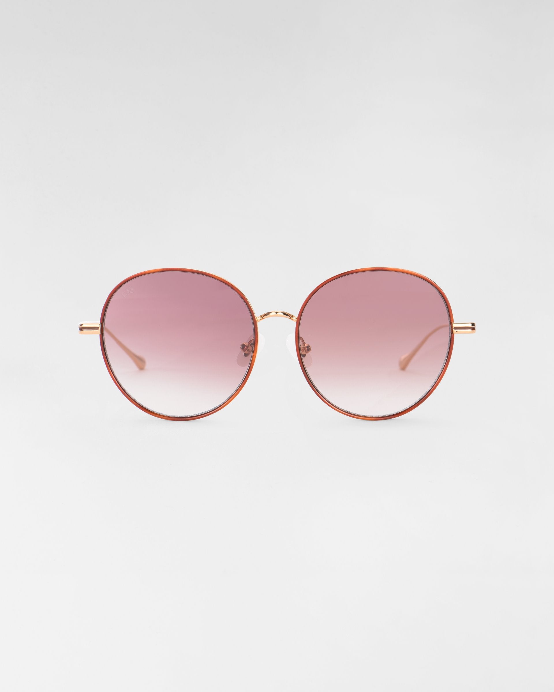 A pair of round Lemon sunglasses from For Art&#39;s Sake® with thin, metallic golden frames and pink-tinted gradient lenses, complemented by jadestone nose pads and 18-karat gold plating, set against a plain, light gray background.