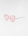 A pair of round, thin-framed For Art's Sake® Lemon sunglasses with pink-tinted lenses and metal temples. The clear nose pads are complemented by hand-painted enamel details. The frame is delicate and minimalistic, set against a plain white background.