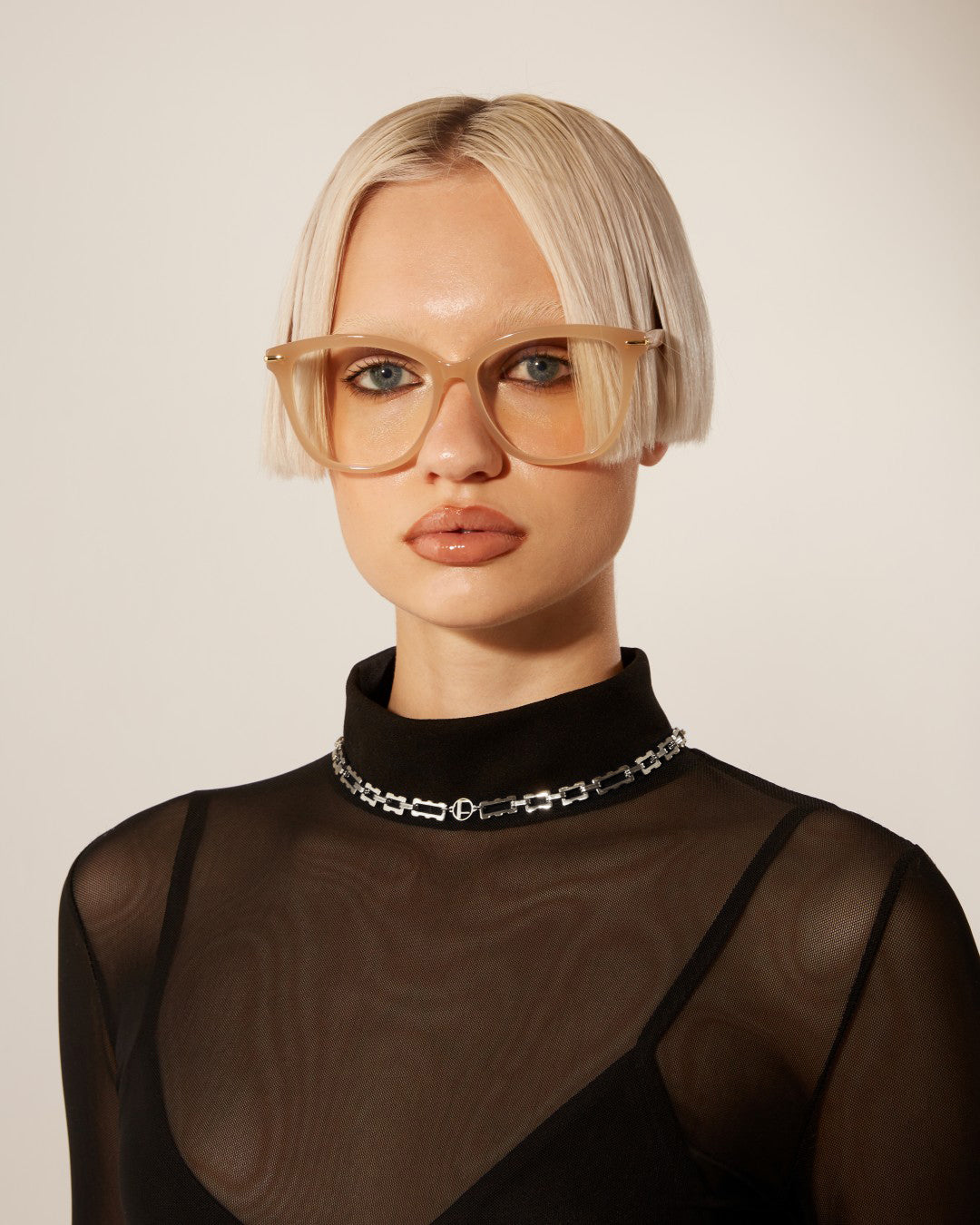 A person with short, platinum blonde hair and blunt bangs wears large, beige glasses and a sheer black top with a high collar. They sport the For Art&#39;s Sake® Links Necklace Silver, adding a touch of elegance to their neutral expression. The background is plain and light-colored.