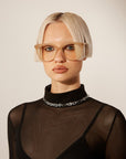 A person with short, platinum blonde hair and blunt bangs wears large, beige glasses and a sheer black top with a high collar. They sport the For Art's Sake® Links Necklace Silver, adding a touch of elegance to their neutral expression. The background is plain and light-colored.