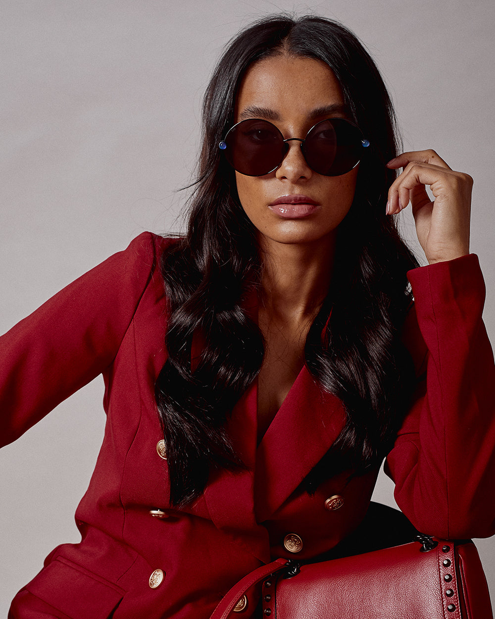 A person with long dark hair wearing round For Art&#39;s Sake® Love Story shades and a red blazer with gold buttons poses confidently against a neutral background. They have a serious expression, and their sunglasses feature jadestone nose pads for extra comfort while offering 100% UV protection. One hand is resting near their temple.