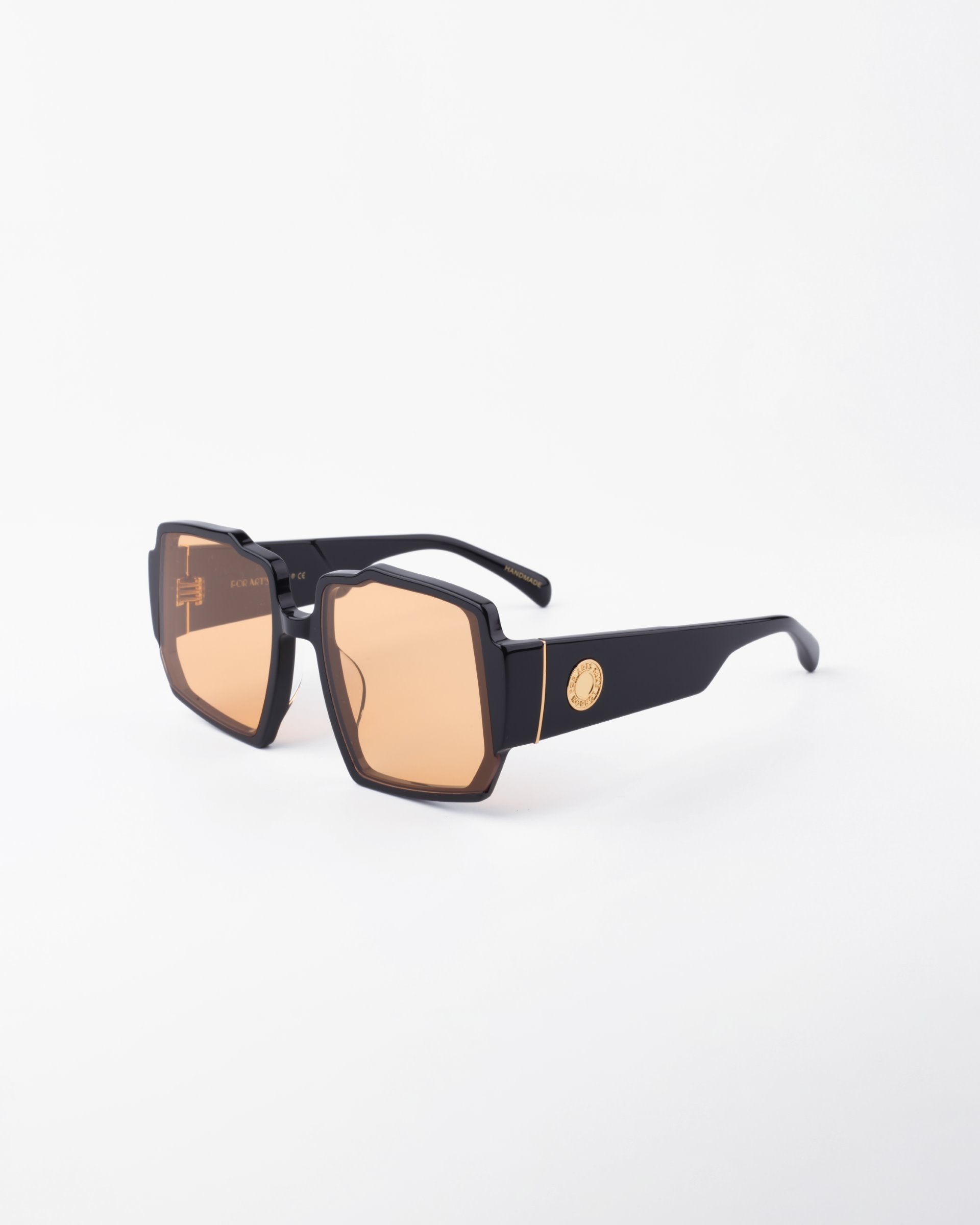 A pair of stylish black For Art&#39;s Sake® Moritz sunglasses with large, geometric, orange-tinted UV-protected lenses sits against a plain white background. The sunglasses feature a chunky acetate frame and thick arms adorned with a small, gold-colored emblem near the hinge.