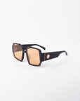 A pair of stylish black For Art's Sake® Moritz sunglasses with large, geometric, orange-tinted UV-protected lenses sits against a plain white background. The sunglasses feature a chunky acetate frame and thick arms adorned with a small, gold-colored emblem near the hinge.
