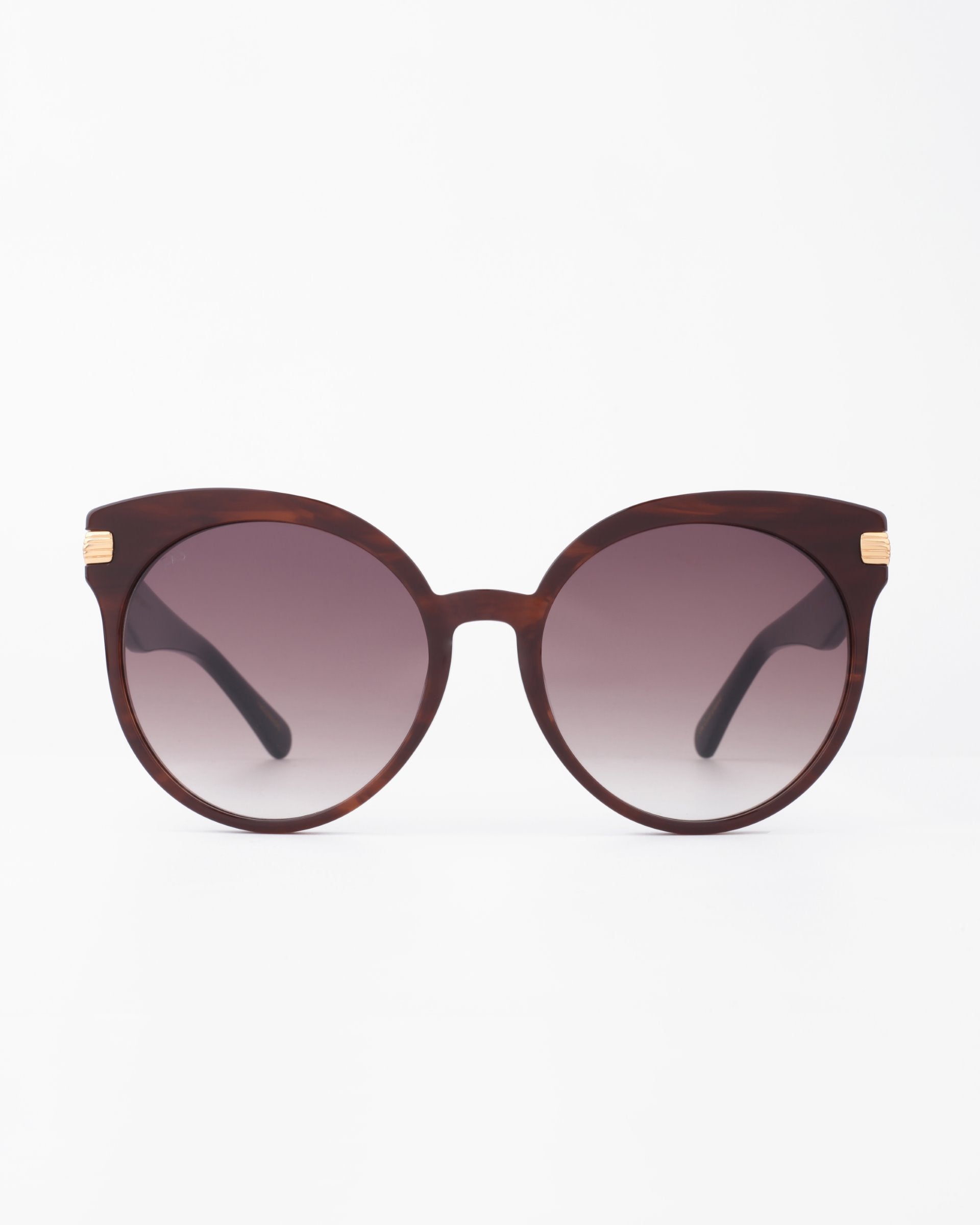 The Muse by For Art&#39;s Sake® is a pair of round brown sunglasses with gradient lenses, featuring 18-karat gold-plated temples. Crafted from handmade plant-based acetate, the Muse sunglasses are placed on a white surface with a minimalistic background.