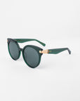 A pair of stylish Muse sunglasses by For Art's Sake® with dark green frames and shatter-resistant nylon lenses is displayed on a white background. The temples feature 18-karat gold-plated detailing near the hinges, adding a touch of elegance to the handmade plant-based acetate design.