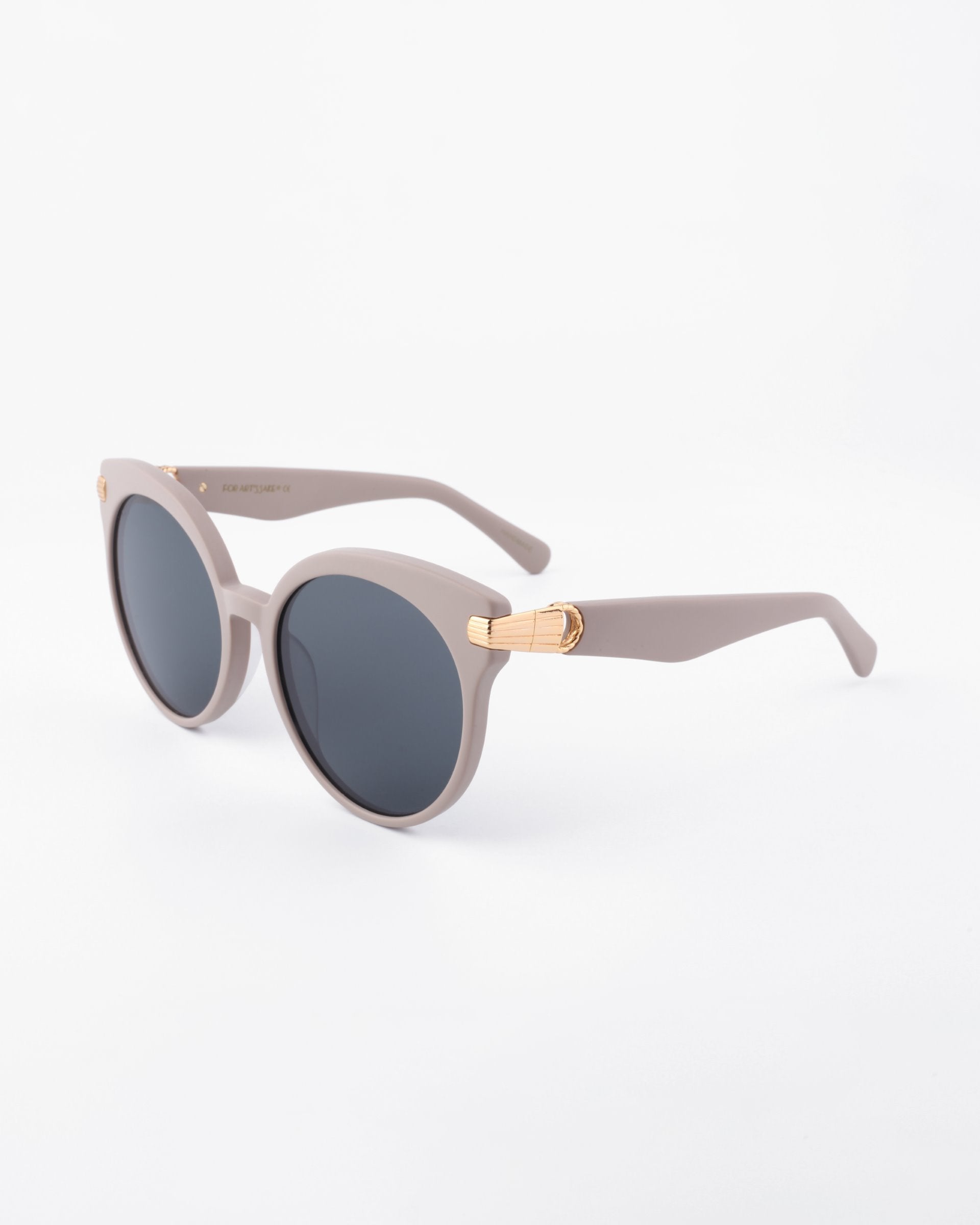 A pair of stylish round For Art&#39;s Sake® Muse sunglasses with light grey frames and shatter-resistant nylon lenses. The arms feature 18-karat gold-plated accents near the hinges. Handmade from plant-based acetate, these sunglasses are placed against a plain white background.