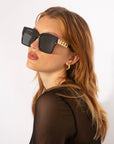 A woman with light brown hair is wearing oversized square frames featuring shatter-resistant nylon lenses and an 18-karat gold plated chain detail on the temples. She is looking over her shoulder, wearing a black sheer top and gold hoop earrings against a plain white background. She is wearing Castle by For Art's Sake®.