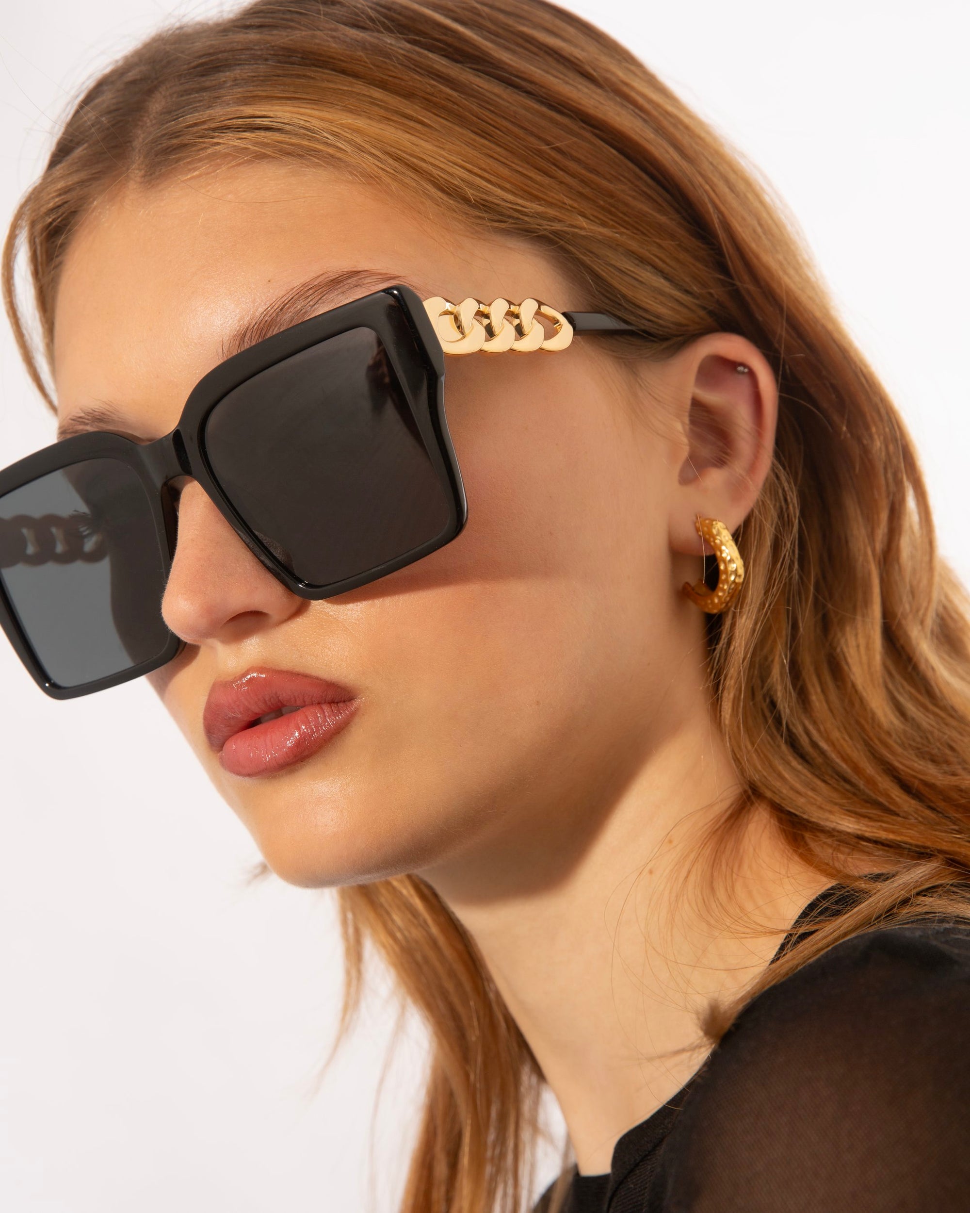 A woman with long, blonde hair wears oversized square-shaped black For Art&#39;s Sake® Castle sunglasses featuring an 18-karat gold plated chain on the frames. She is also wearing small gold hoop earrings and a black top. The background is plain white.