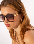 A woman with long, light brown hair is wearing oversized square frames with ultra-lightweight shatter-resistant lenses and decorative gold accents on the temples from For Art's Sake® called Castle. She is also wearing 18-karat gold plated hoop earrings and a thin gold chain necklace. The background is plain and white.