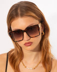 A person with shoulder-length auburn hair wears large, square, tortoiseshell For Art's Sake® Castle sunglasses featuring ultra-lightweight shatter-resistant lenses, gold hoop earrings, and a gold necklace with pearls. The background is plain white, and the person is looking slightly down.