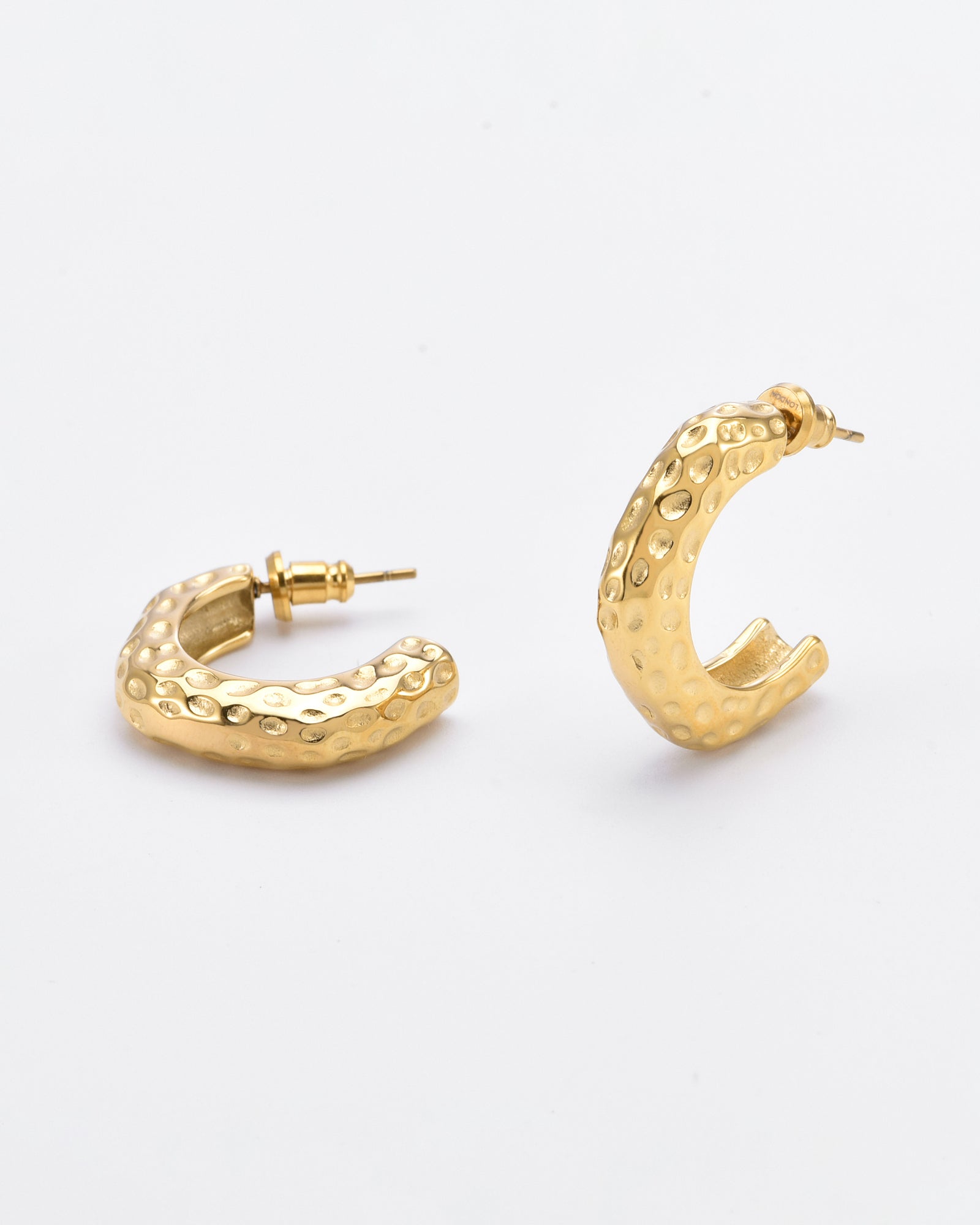 A pair of For Art's Sake® Mia Earrings Gold are shown against a plain white background. The gold-plated earrings boast a textured surface with a hammered appearance, featuring subtle dimples all over. One earring is standing upright, while the other lies flat.