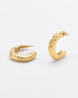 A pair of For Art's Sake® Mia Earrings Gold are shown against a plain white background. The gold-plated earrings boast a textured surface with a hammered appearance, featuring subtle dimples all over. One earring is standing upright, while the other lies flat.